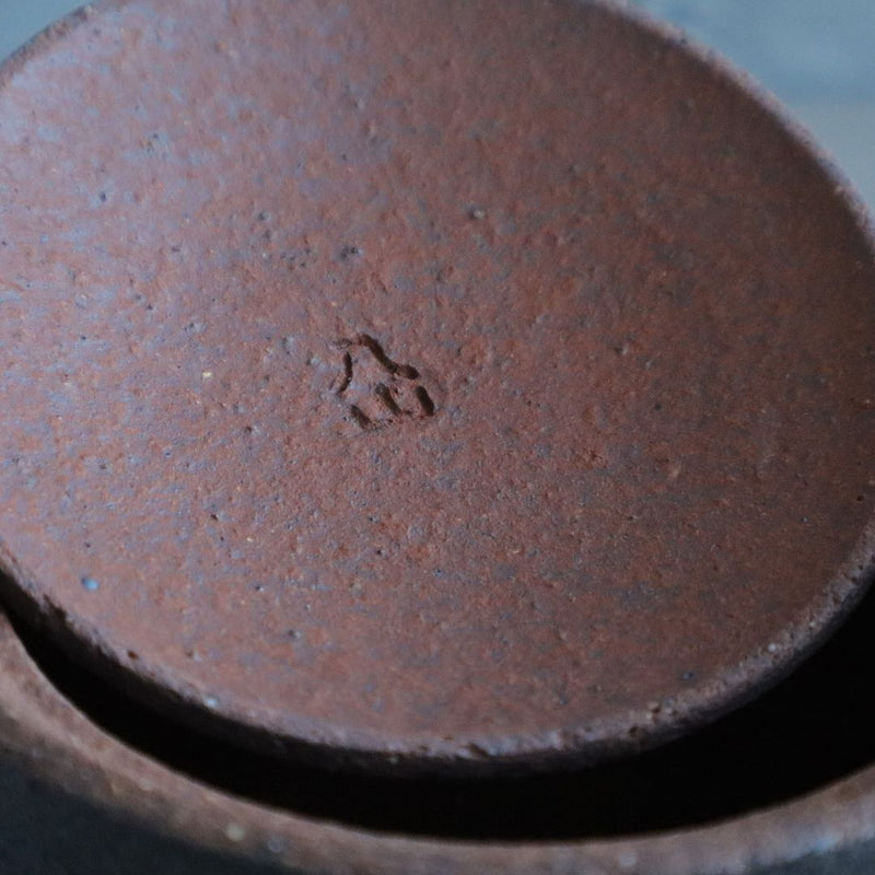 A detailed view of the top of a black ceramic salt cellar with its textured brown lid slightly ajar, revealing a maker& mark impressed into the clay.