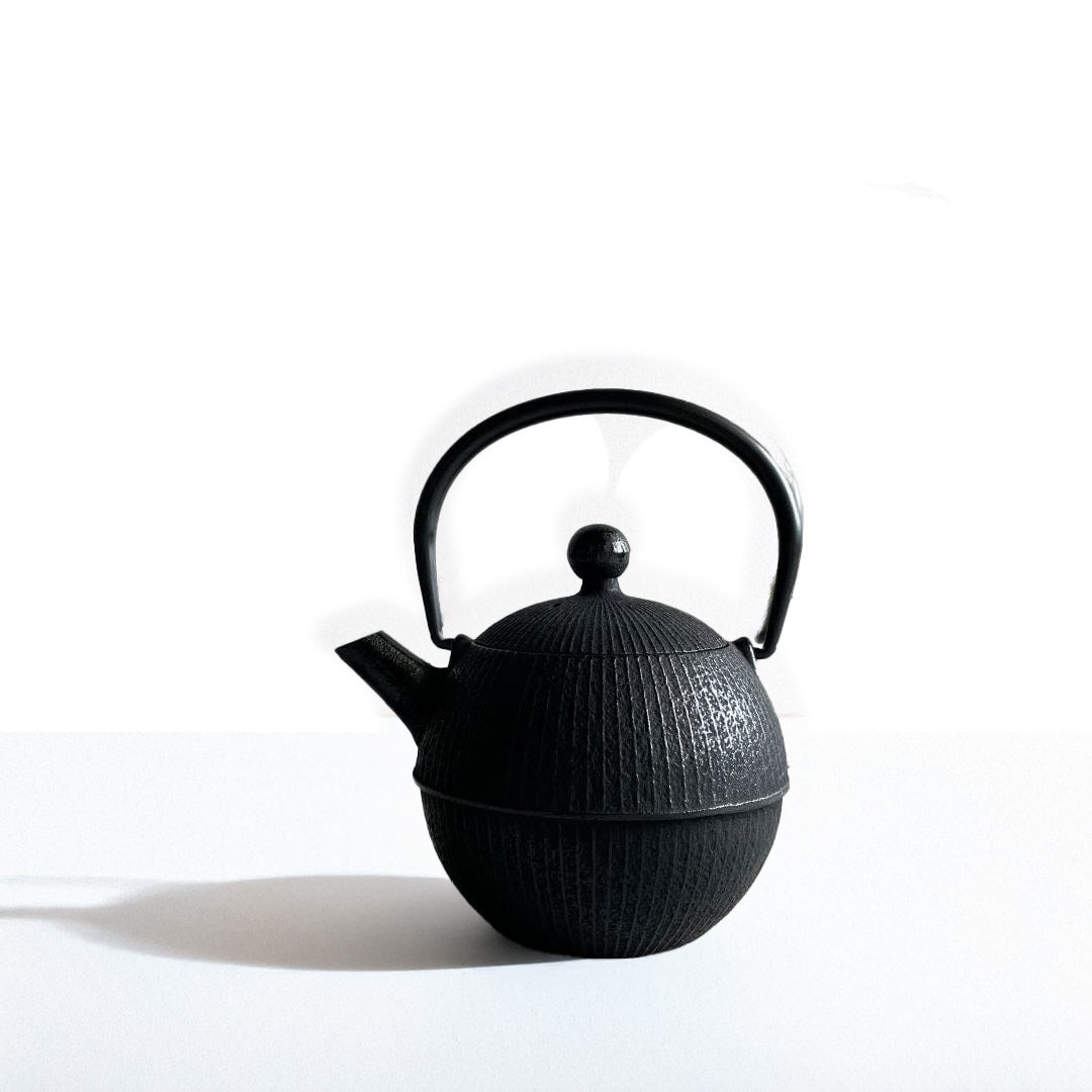 This is a front view of a black cast iron Tetsubin teapot with a lightly rough surface on the body. The teapot has a round shape with vertical stripes all around the surface. The iron handle has a smooth surface and the teapot is standing in the middle of a space with a grey wall and grey surface.