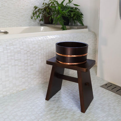 A black wooden stool with a sleek design holds a black bucket with copper bands, both crafted from Japanese Hinoki Cypress, set against a white tiled bathroom with a green plant in the background.