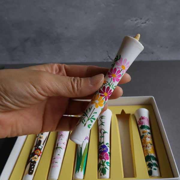 A hand holds a wax candle adorned with a vibrant floral design, above a box showcasing a variety of similarly decorated candles, all set against a grey textured background.