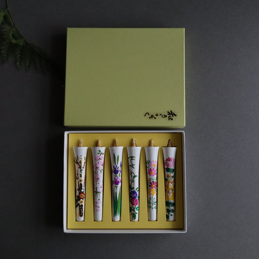 A set of six hand-painted rice bran wax candles with seasonal floral designs, neatly arranged in a box. Each candle measures 12cm or 4.7 inches in length. The box is olive green with Japanese characters on the lid, presented on a dark surface.