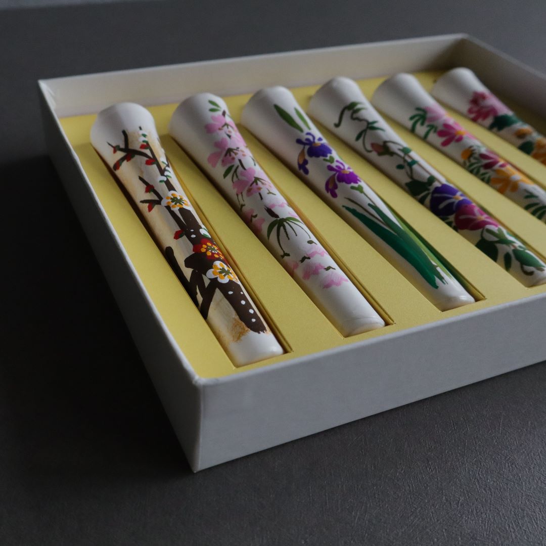 A close-up, angled view of six hand-painted wax candles with floral designs, arrayed in a box with a yellow interior, against a grey backdrop with an evergreen branch visible.