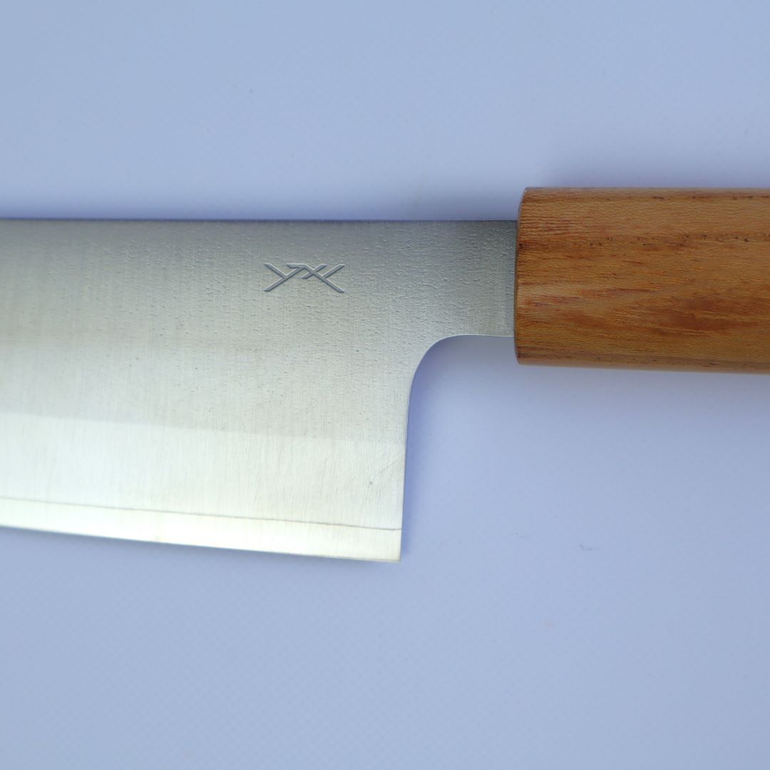 Close-up of a Santoku knife's blade and wooden handle against a light background. The blade has a brushed metal finish with a distinctive maker's mark near the handle, highlighting the blade's sharp edge and craftsmanship. The wooden handle has a rich, natural grain, suggesting quality and durability.