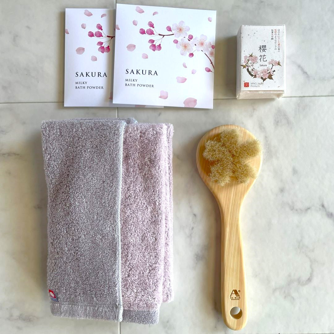 Japanese Sakura Bath Gift Set displayed on a marble surface, featuring an Imabari reversible towel, a natural wooden body brush, Sakura soap, and two sachets of Sakura milky bath powder with cherry blossom packaging.