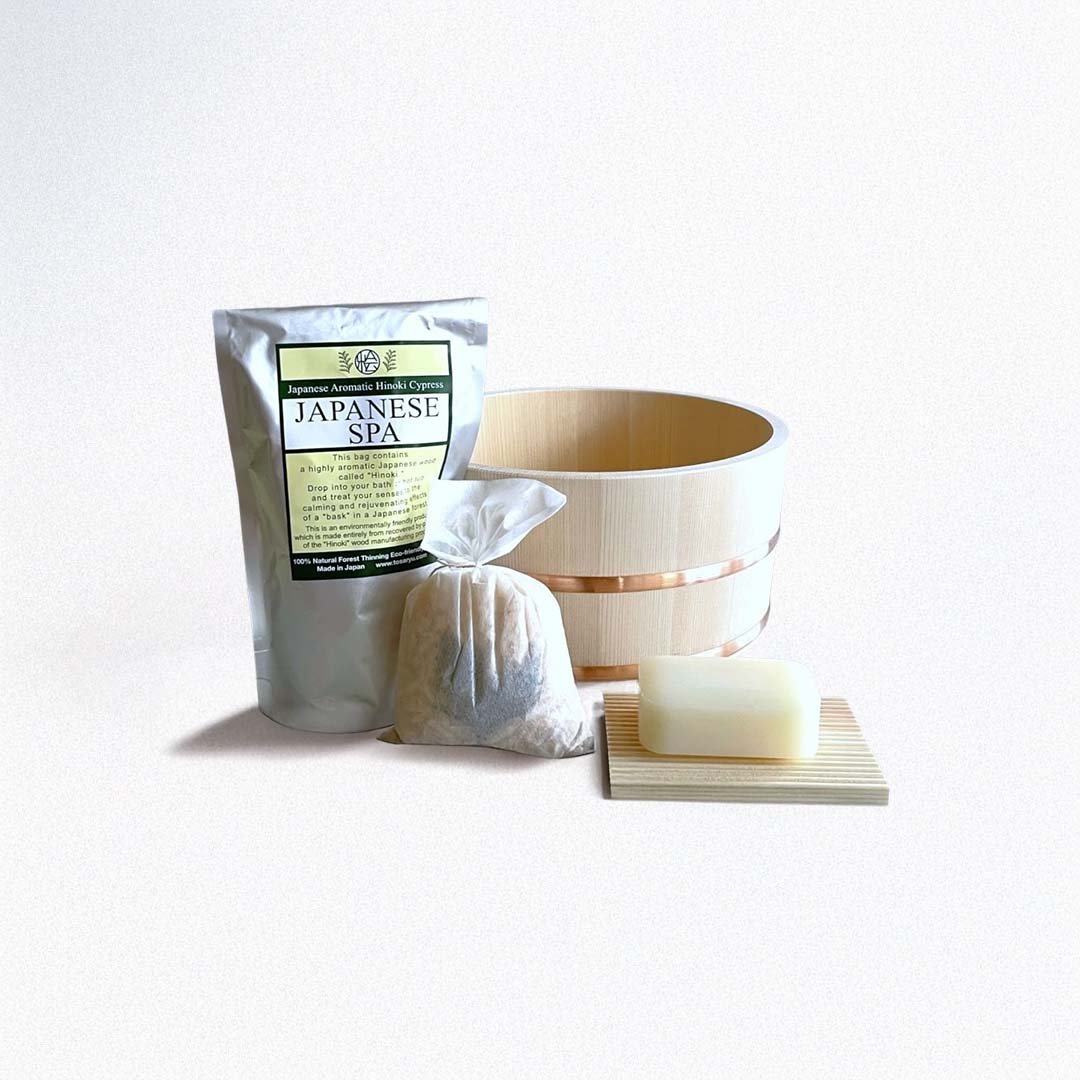 A spa set is displayed on a concrete surface against a concrete wall. The set includes a light-colored wooden bucket with a copper band, a white package labeled "Japanese Aromatic Hinoki Cypress JAPANESE SPA," a white cloth bag with contents creating a rounded shape, and a piece of soap resting on a light wood soap dish with ridges