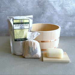 A spa set is displayed on a concrete surface against a concrete wall. The set includes a light-colored wooden bucket with a copper band, a white package labeled "Japanese Aromatic Hinoki Cypress JAPANESE SPA," a white cloth bag with contents creating a rounded shape, and a piece of soap resting on a light wood soap dish with ridges. The overall aesthetic suggests a serene, minimalistic spa experience with an emphasis on natural Japanese elements.