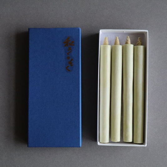 An open box of organic sumac wax taper candles lies next to its blue lid with Japanese characters, on a gray background. The natural-colored candles are neatly arranged inside the white box, ready for display or use.