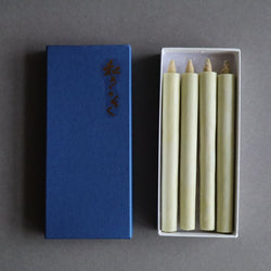 An open box of organic sumac wax taper candles lies next to its blue lid with Japanese characters, on a gray background. The natural-colored candles are neatly arranged inside the white box, ready for display or use.