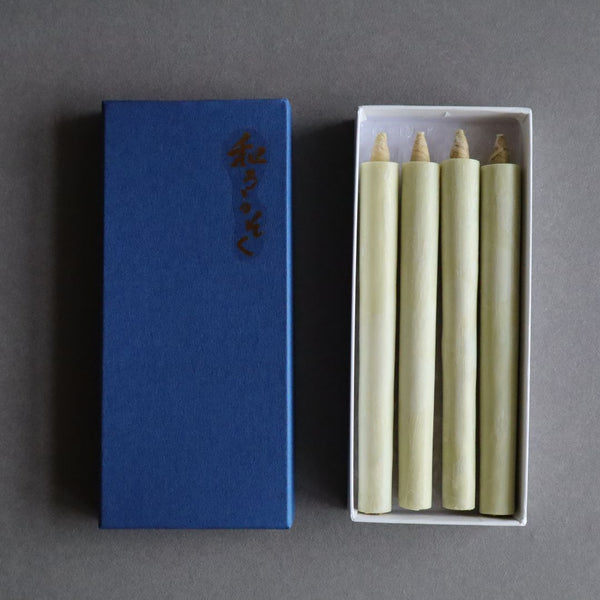 An open box of organic sumac wax taper candles lies next to its blue lid with Japanese characters, on a gray background. The natural-colored candles are neatly arranged inside the white box, ready for display or use.