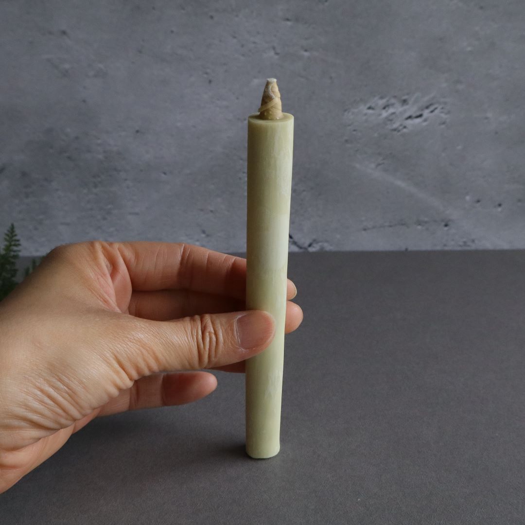 A hand is holding a single Japanese sumac wax taper candle against a textured gray background. The candle, slender and pale, with a pointed wick, appears to be around 11cm (4.3 inches) long. In the corner, a white box is partially visible.