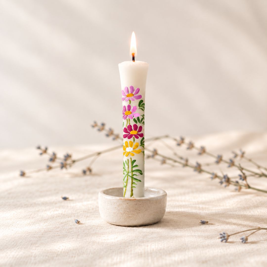 A white taper candle with colorful hand-painted flowers stands lit in a small round ceramic holder on a beige fabric surface with dried floral stems in the background.