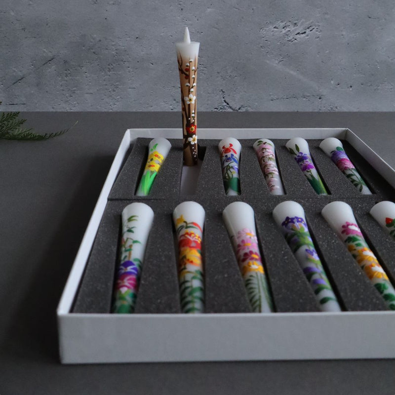 A single lit hand-painted candle stands out, elegantly poised before a grey textured backdrop, with an array of unlit, vibrantly designed sumac wax candles in a box below, showcasing an artistic contrast and the gentle allure of candlelight.