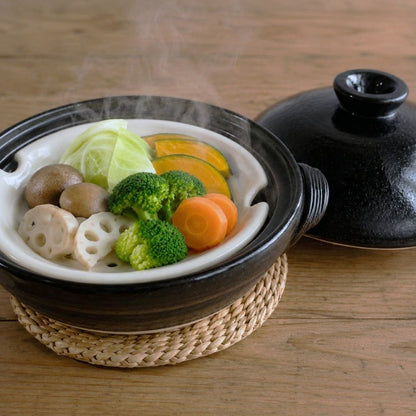 A black ceramic steaming pot with a white interior, filled with steaming vegetables including broccoli, carrots, mushrooms, and lotus slices, placed on a woven mat on a wooden table. The lid rests beside the pot.