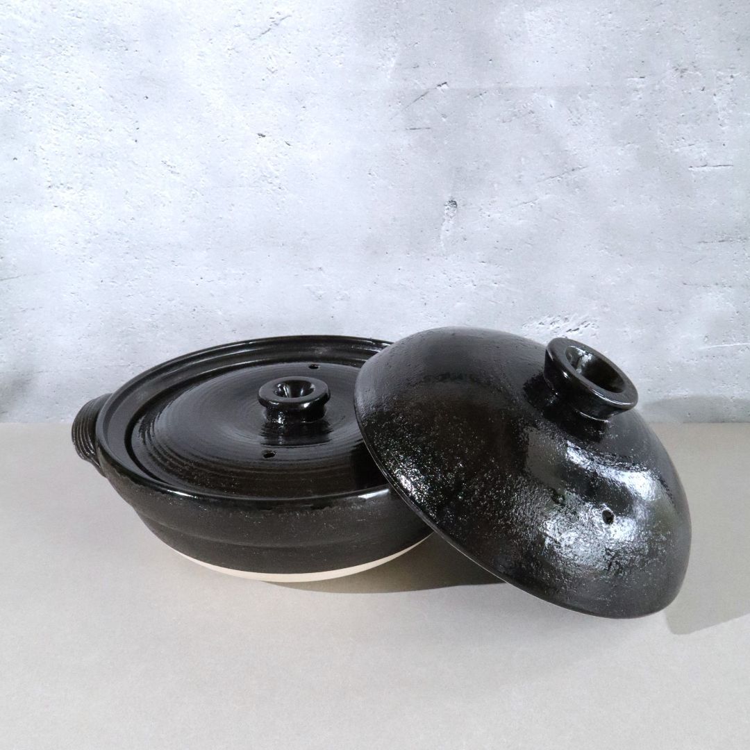 A black glazed ceramic lidded casserole dish with a matching lid leaning against it, set on a light surface against a textured light gray wall.