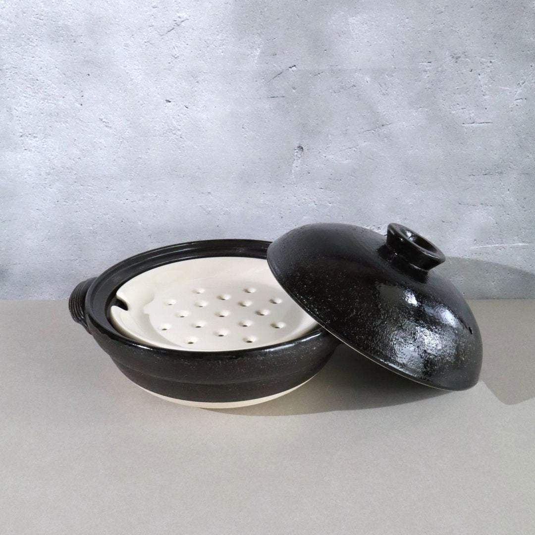 A black glazed ceramic pot with its lid leaning against it, showing a white perforated insert inside. The pot has a side handle and sits on a light gray surface against a textured gray wall.