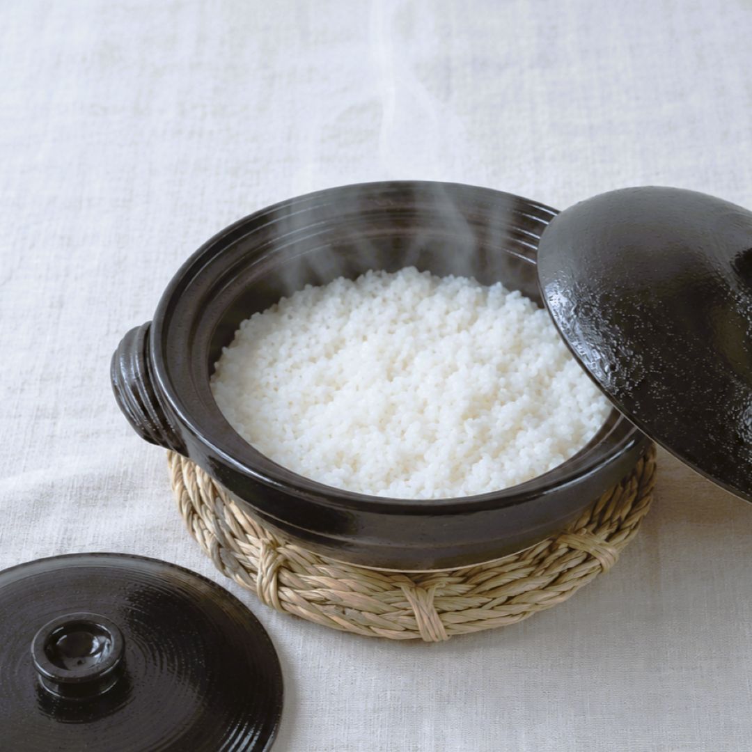 A dark ceramic rice cooker pot filled with steaming white rice, set on a woven straw base with one lid leaning against it and another lid placed beside it on a light fabric surface.