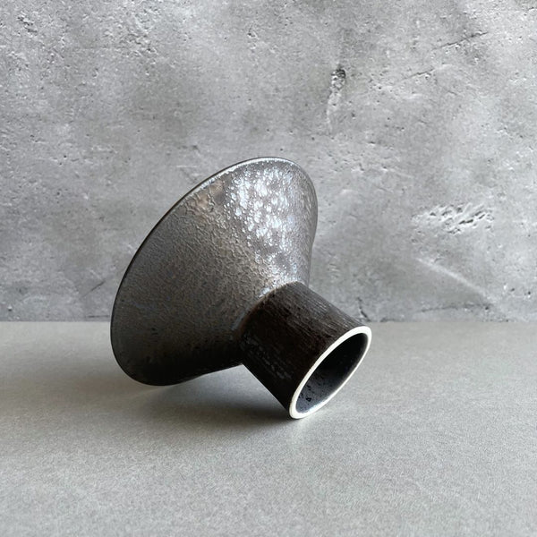 A round black ceramic plate with a foot, shown fallen on a grey table.