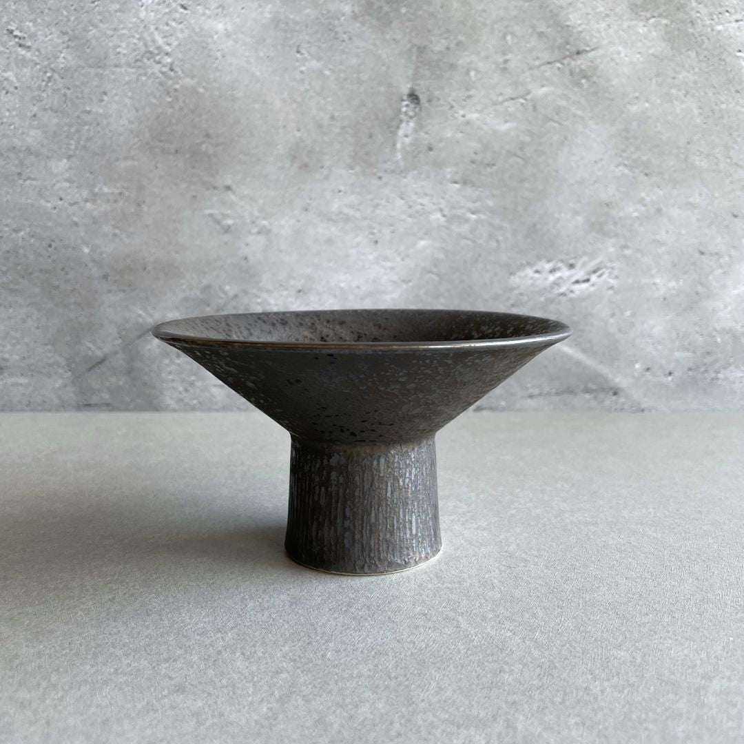 A side view of a round black ceramic plate with a visible foot, placed on a grey table.