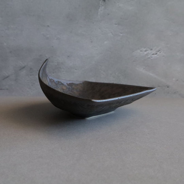 Dynamic side view of a uniquely shaped Japanese porcelain tray resting on a gray surface. The tray dramatic angular form elevates one corner high, showcasing the metallic glaze with intricate mottled patterns in shades of gray, black, and blue.