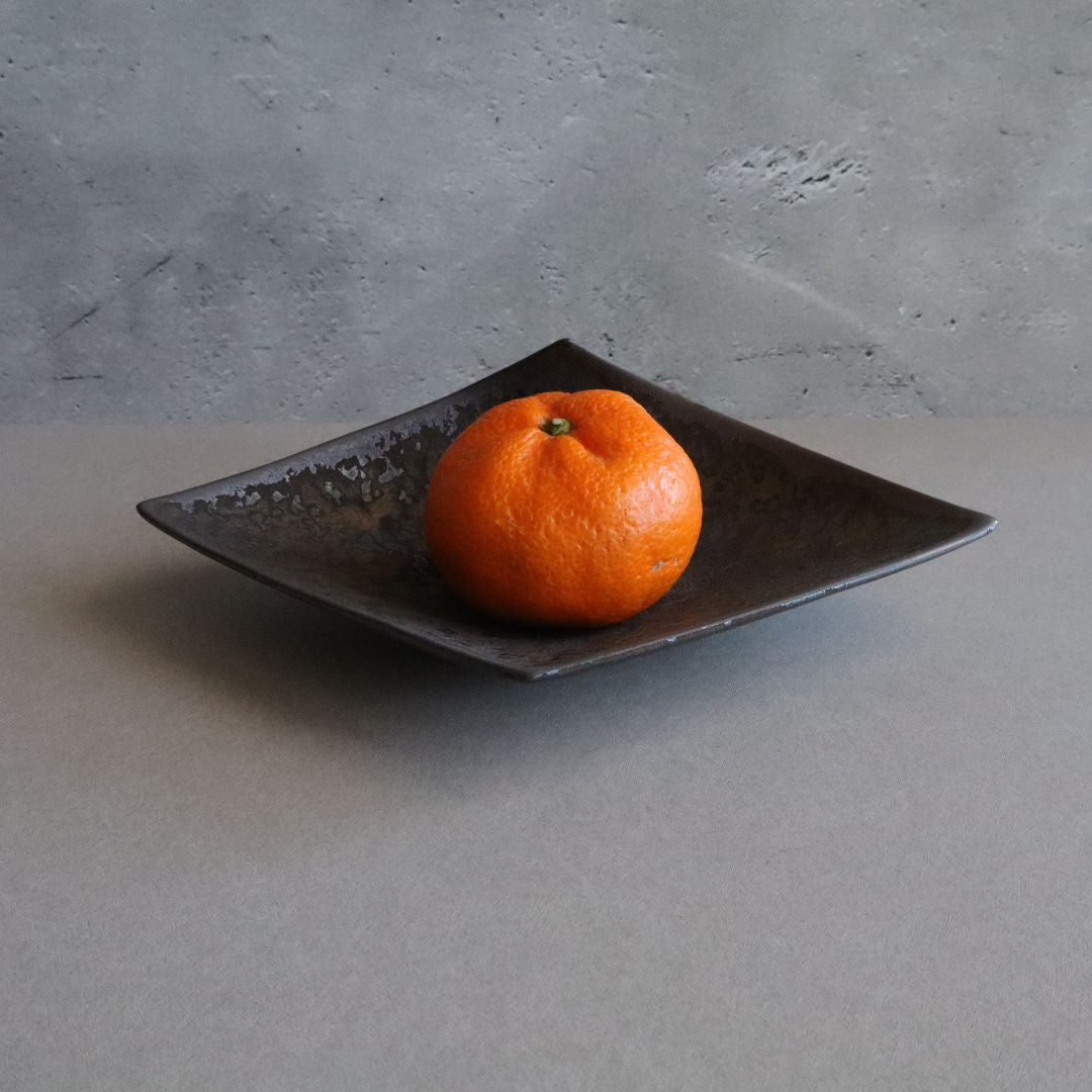A vibrant orange mandarin rests on a dark, handcrafted Japanese porcelain tray. The tray surface is adorned with a metallic glaze, featuring mottled shades of black and gray.