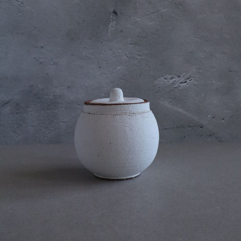 A white handmade Japanese Shigaraki ceramic salt cellar with a rustic brown-edged lid, centered on a grey textured background.