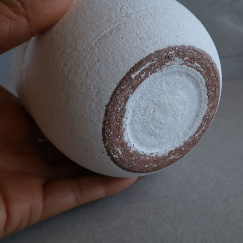 A close-up view of a white handmade Japanese Shigaraki ceramic container held in a hand, showcasing the brown-edged circular opening at the base against a gray background.