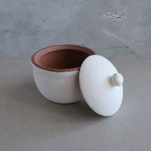 A handmade white Shigaraki ceramic container with its lid tilted to the side, revealing the unglazed terra cotta interior. The lid, with a small central knob, matches the container exterior, except for the brown-trimmed edge.