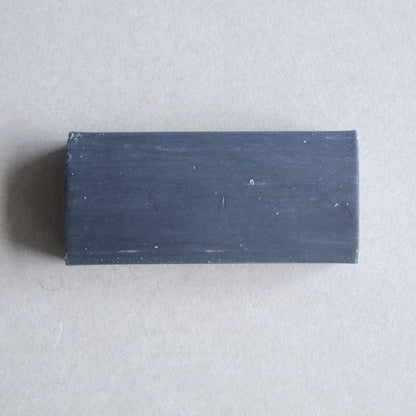 A rectangular bar of black charcoal facial soap with a smooth, matte finish, placed on a neutral beige surface. The soap has a minimalist design with slight imperfections on the surface, highlighting its natural and handcrafted quality.