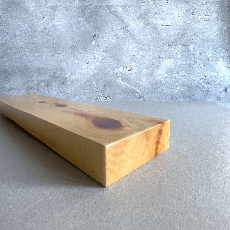 A close-up view of a pine wooden shelf with a natural finish, showcasing its smooth surface and the detailed wood grain and knots. The shelf is angled upwards against a rough, grey concrete wall, emphasizing the wood's warm tones and textures.