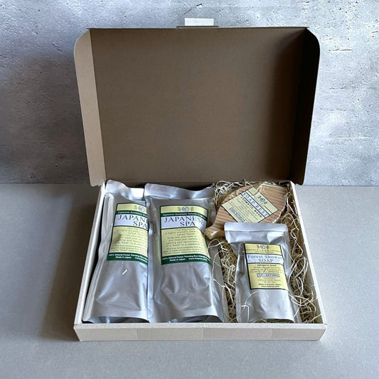 A pack of spa accessories in a grey room, featuring two spa bags filled with hinoki flakes, a wooden soap dish, and a soap infused with hinoki leaf essential oil.