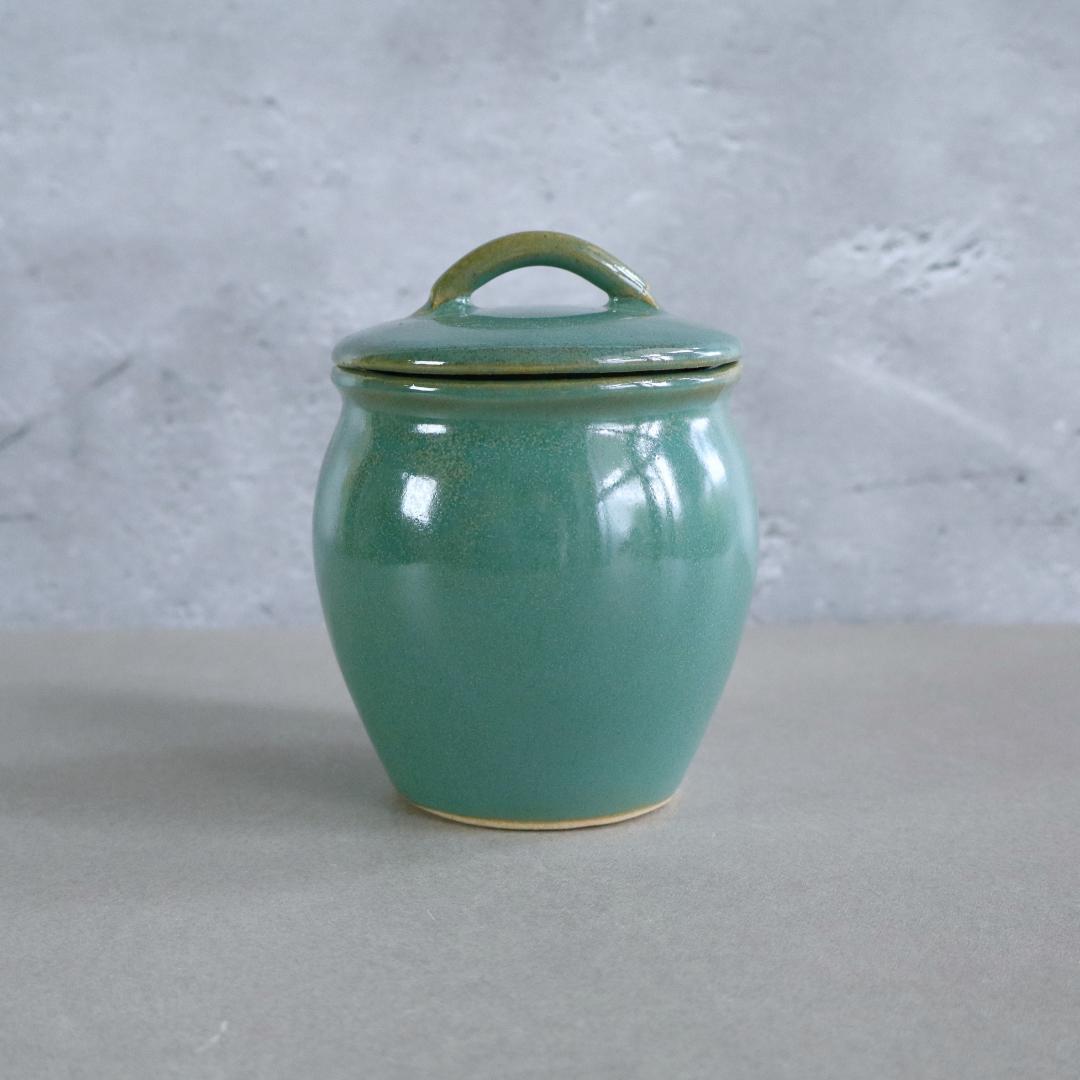 Handmade Japanese ceramic jar with a lid, featuring a smooth retro green glaze. This Mashiko Ware jar has a curved silhouette and a convenient handle on the lid, making it ideal for storing spices, tea, or small keepsakes.