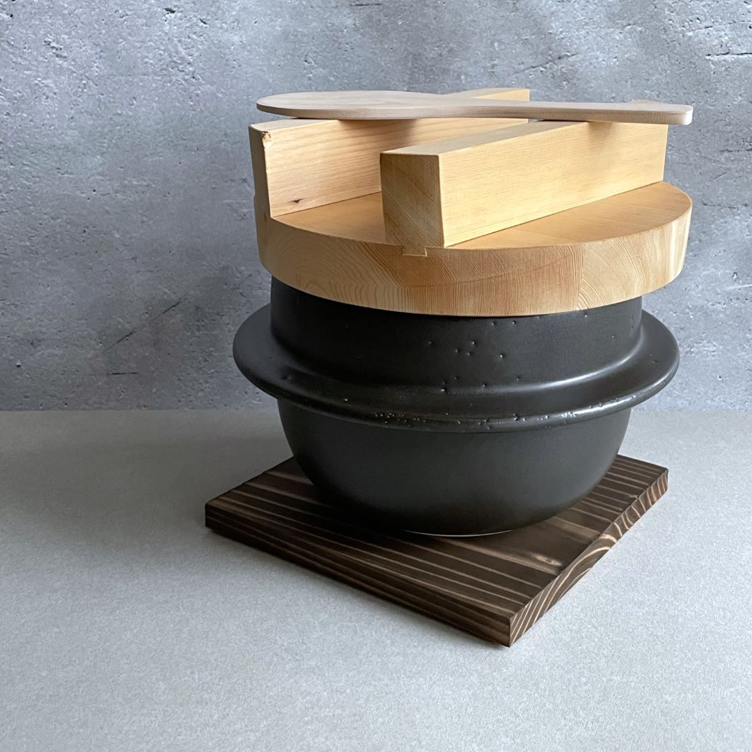 A Japanese handmade rice cooker with a wooden lid, standing in front of a grey wall, featuring a black clay pot, and a wooden spatula resting on top of the wooden lid.