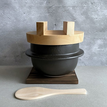 A Japanese handmade rice cooker with a wooden lid, placed in front of a grey wall, accompanied by a black clay pot and a wooden spatula.