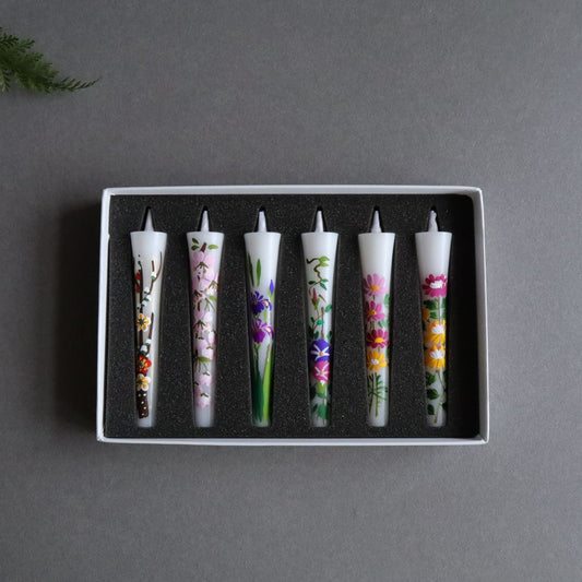 A set of six hand-painted sumac wax candles, each with unique floral designs, arranged in a box on a grey surface with a fern leaf accent, each candle measuring 10cm/4 inches.