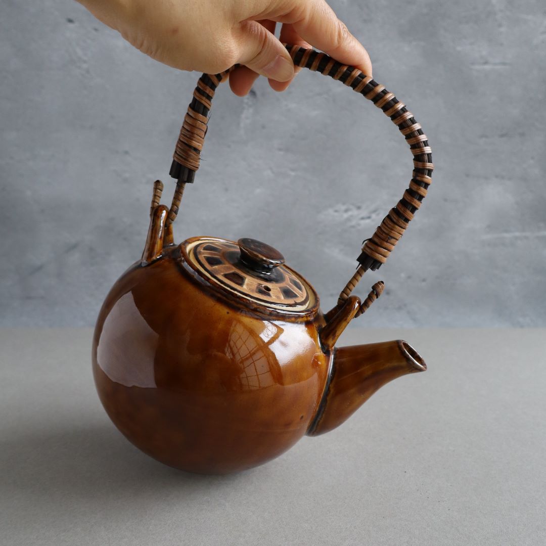 A hand lifting a handmade Japanese ceramic teapot by its woven bamboo handle. The teapot features a glossy brown glaze, a traditional Kyusu-style spout, and an intricately designed lid, showcasing authentic Japanese craftsmanship.