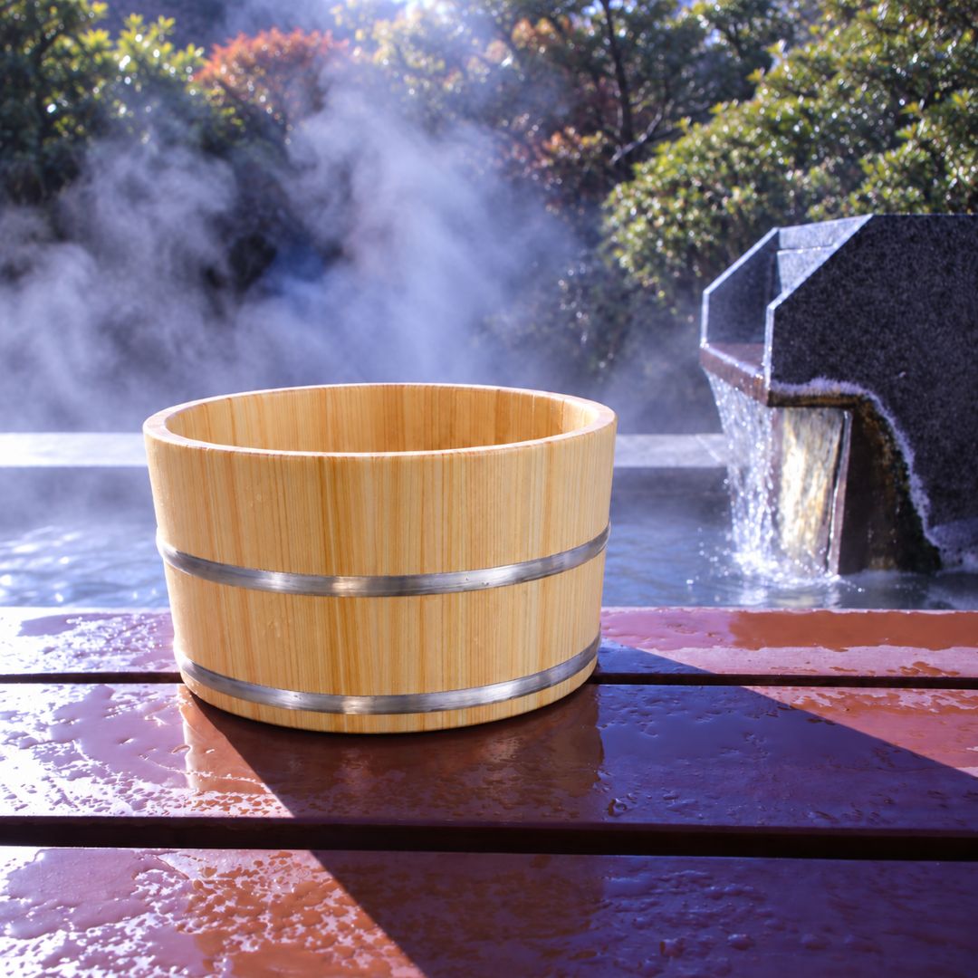 A light natural bamboo hot spring bath bucket with metal bands, sitting on a wooden deck at an outdoor Japanese onsen with steam rising and water flowing in the background.