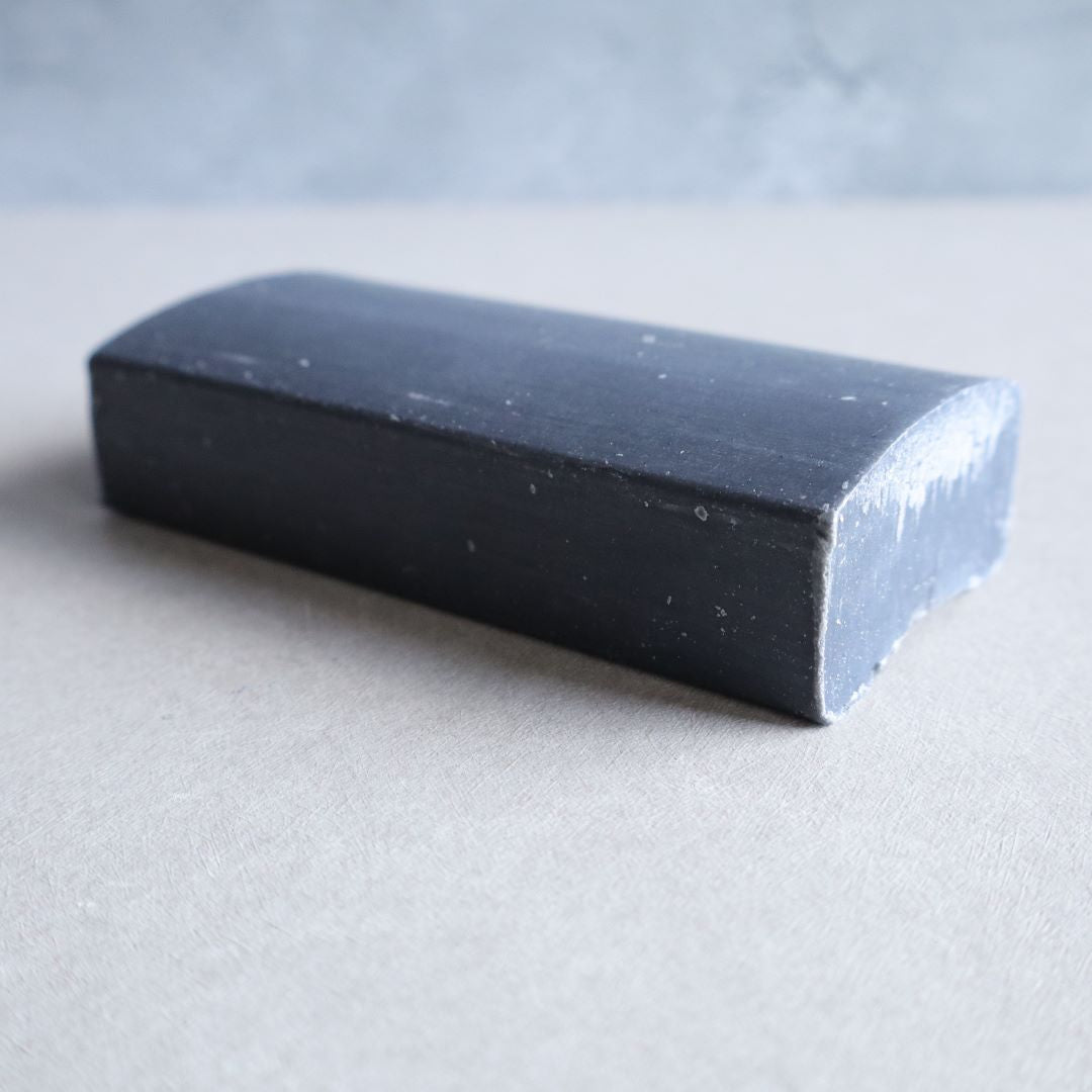 A black charcoal facial soap bar with a smooth, rectangular shape and slightly rounded top edges, placed on a neutral beige surface. The soap has a deep black color with a slightly rough texture on one end, giving it a natural, handcrafted appearance. The background is minimalistic with a soft gray tone.