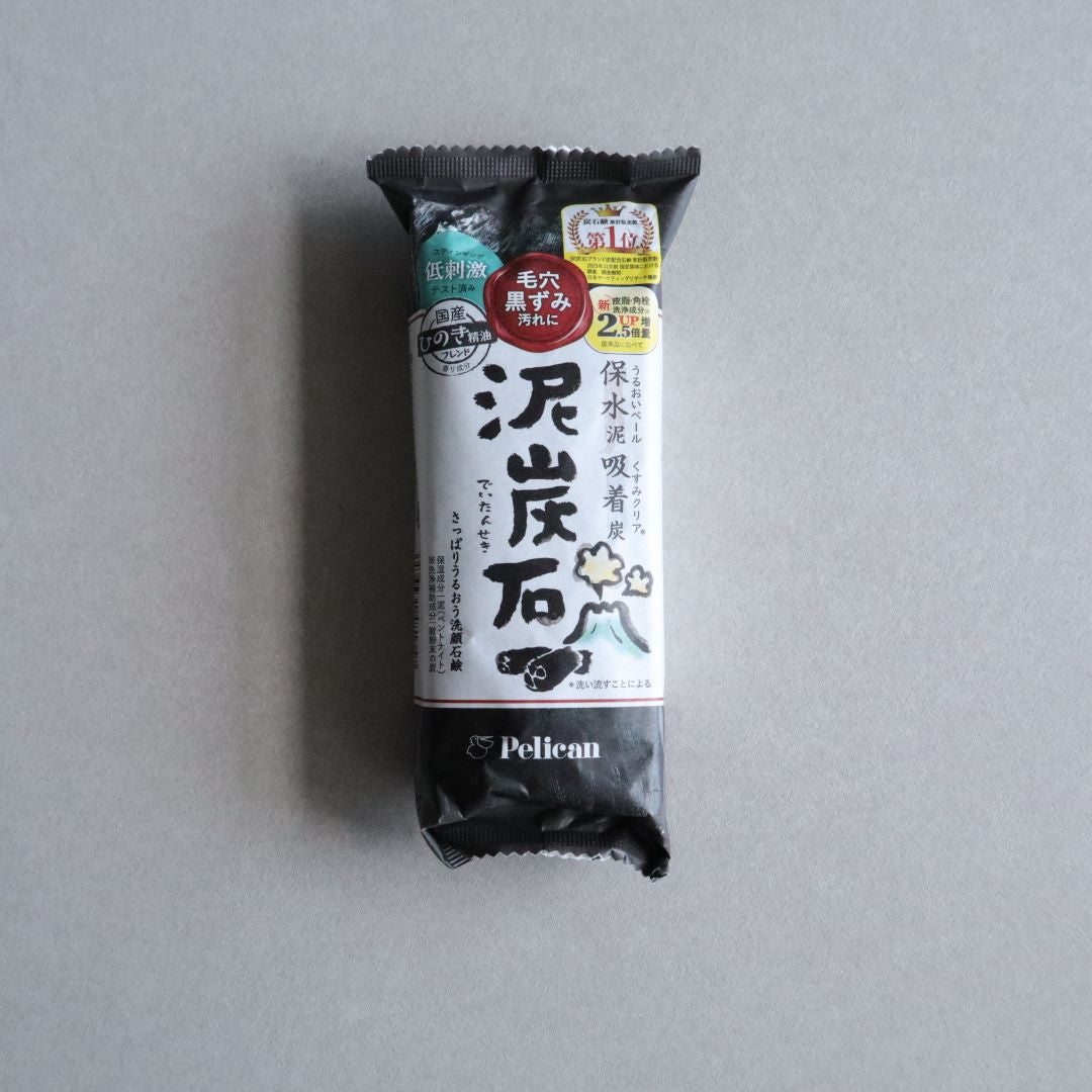 A packaged bar of Japanese Hinoki Mud &amp; Charcoal Facial Soap by Pelican placed on a neutral gray surface. The packaging is primarily black and white with Japanese text, decorative elements, and labels indicating its deep cleansing and moisturizing properties. The design features a charcoal and mud illustration, highlighting its natural skincare benefits.