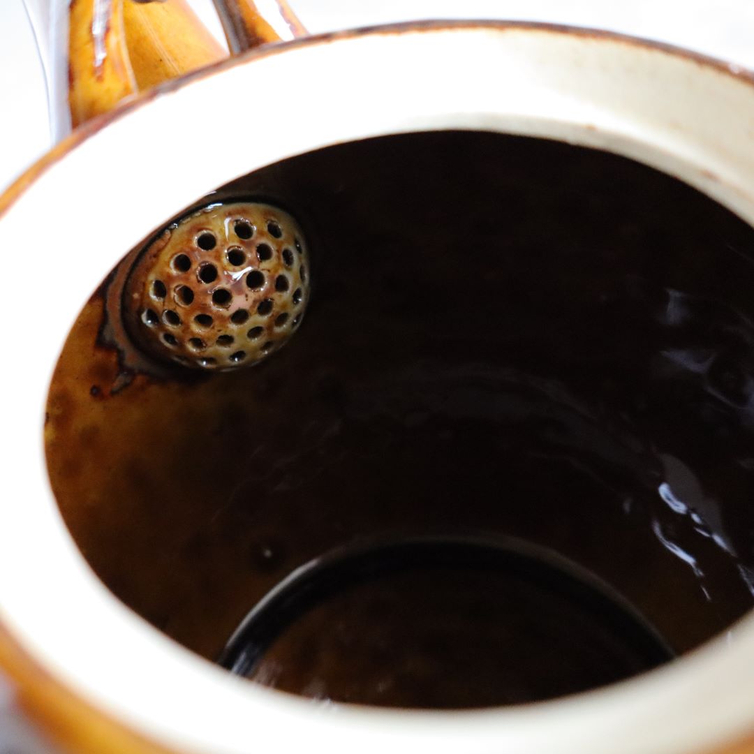 Close-up view of the interior of a handmade Japanese ceramic teapot, showing the built-in ceramic strainer with multiple small holes, designed for brewing loose-leaf tea. The teapot's glossy brown glaze and traditional craftsmanship are visible.