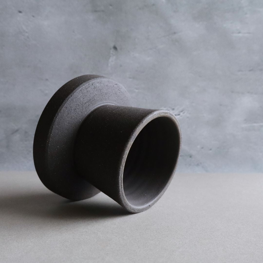 A black footed ceramic bowl is tipped on its side against a textured grey backdrop, showcasing the item's unique shape and the matte finish of the pottery, which emphasizes its modern and minimalist design.