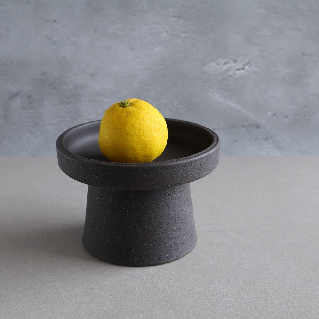 A single bright yellow lemon resting on a minimalist black footed pottery bowl, contrasting against a textured grey background, highlighting the bowl&