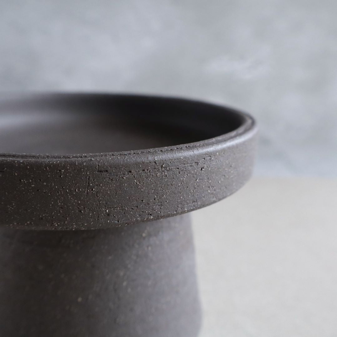  Close-up of the edge of a minimalist black ceramic footed bowl, highlighting the textured surface and the simple yet elegant design of the pottery against a soft-focus grey background.