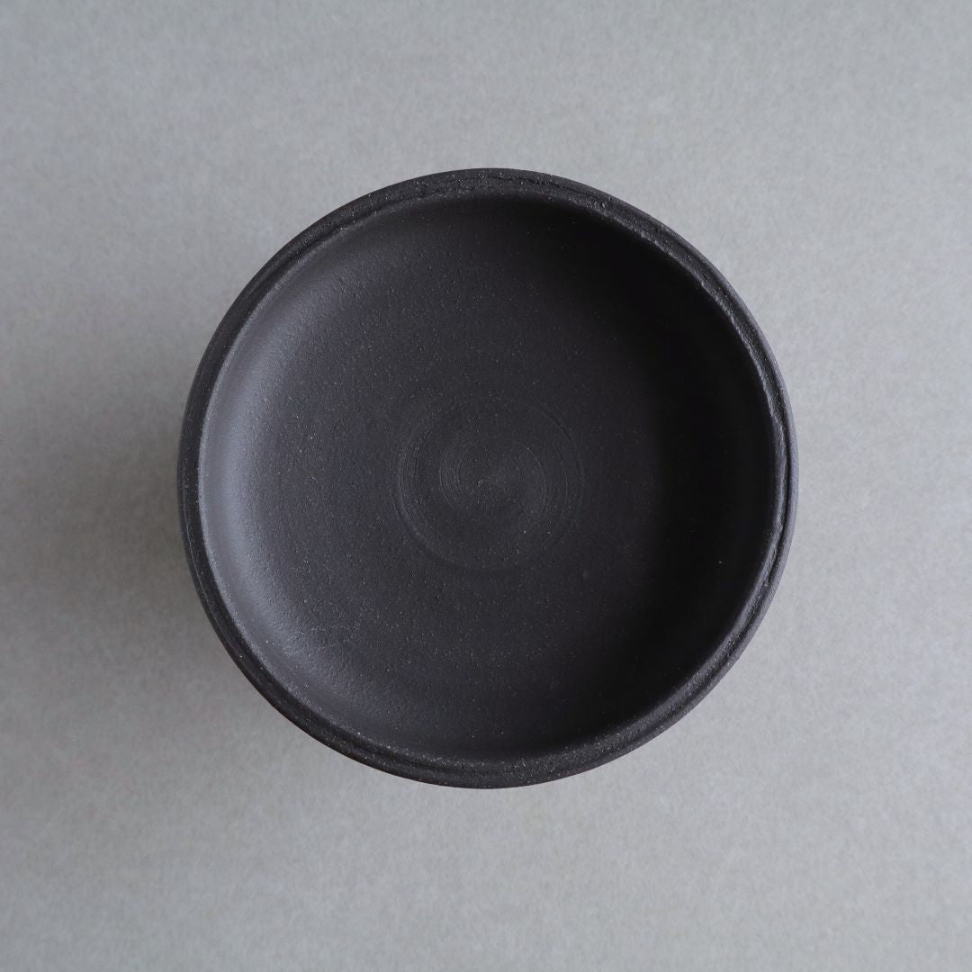 Top-down view of a minimalist black footed bowl, showcasing the circular design inside and the uniform matte finish of the ceramic, set against a neutral grey background.