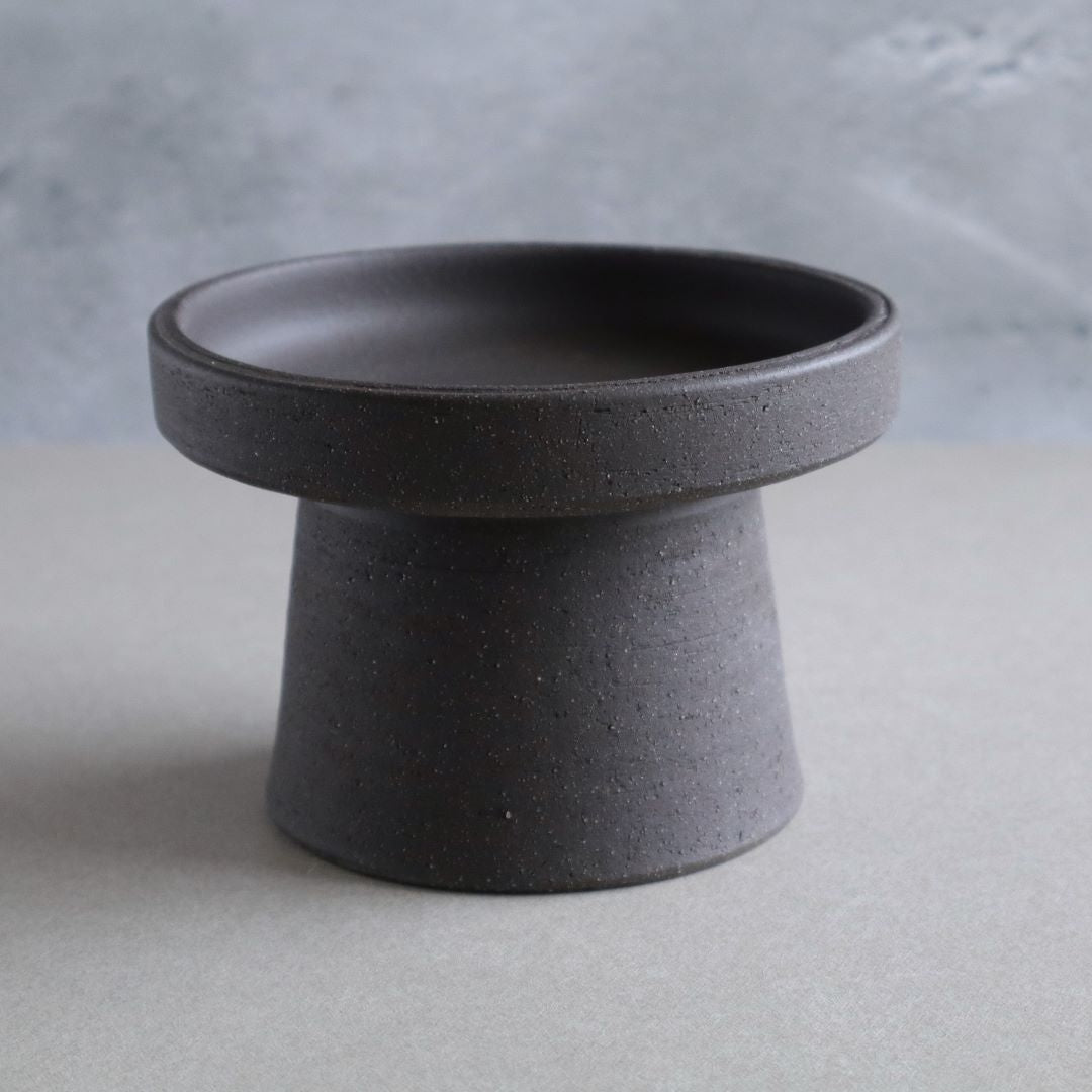 Minimalist black pottery footed bowl on a neutral background, exhibiting a modern take on traditional Japanese Kodai ceramic design, with a textured matte finish.