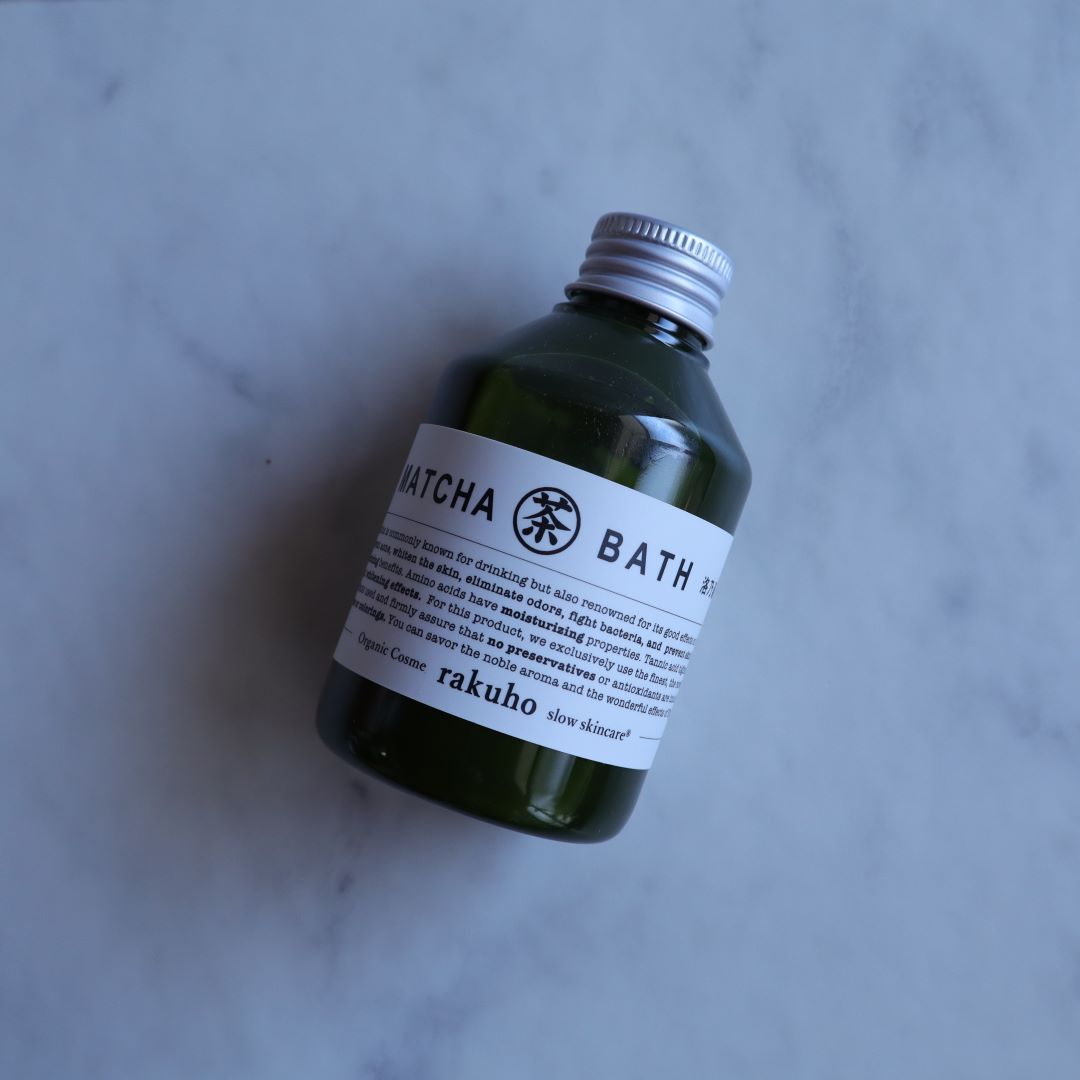A single bottle of Matcha Bath Lotion by &quot;rakuho&quot; rests on a light marble surface. The minimalist white label highlights its organic composition and Japanese craftsmanship, presenting a serene and luxurious skincare aesthetic.