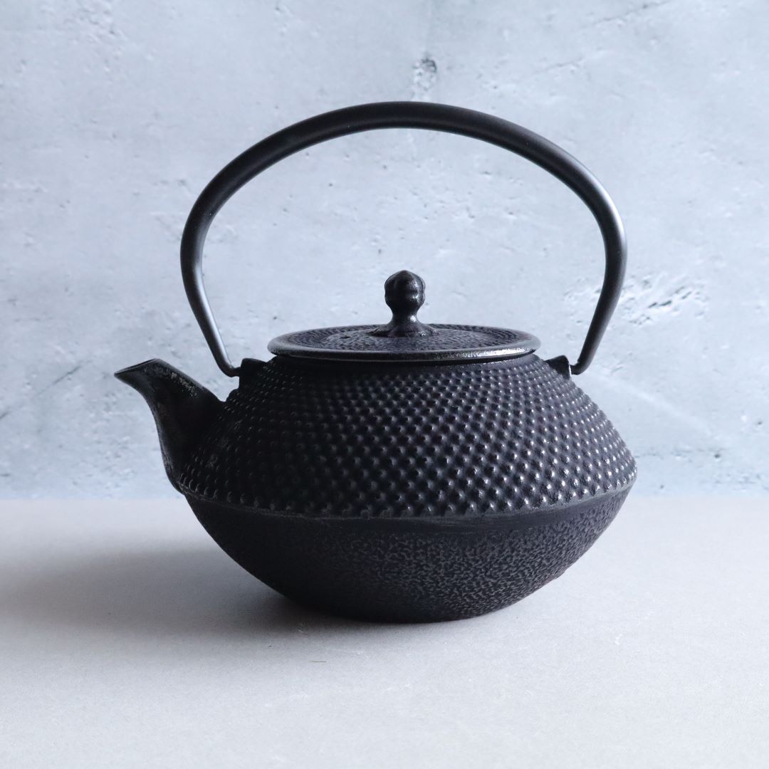 Black cast iron Japanese teapot in Arare Nambu style, featuring a textured dotted surface and curved handle, placed on a light grey surface with a soft concrete background.