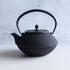 Black cast iron Japanese teapot in Arare Nambu style, featuring a textured dotted surface and curved handle, placed on a light grey surface with a soft concrete background.