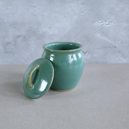 Handmade Japanese ceramic jar with a retro green glaze, shown with its lid removed. This Mashiko Ware jar has a smooth, glossy finish and a curved shape, perfect for storing spices, tea, or small keepsakes.