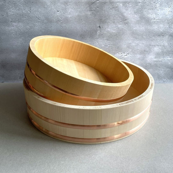 Front view of a brown wooden Hangiri sushi bucket with two copper rings around the rim, placed on a grey surface against a grey background.