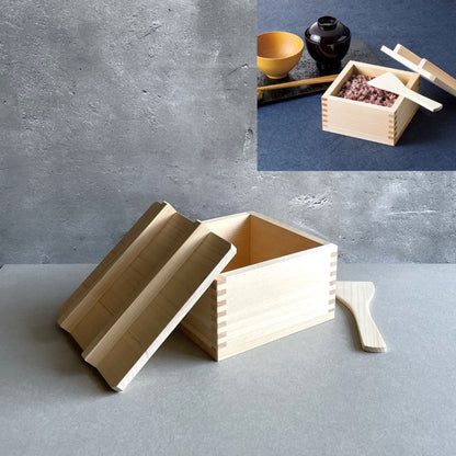on the top, a wooden rice container next to a soup  bowl and under wooden rice container with open lid on side of the box and kitchen tool next to it